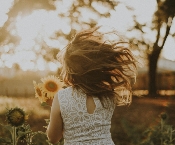 Mädchen im weißen Spitzenkleid mit Sonnenblumen im Haar, von hinten im warmen Abendlicht
