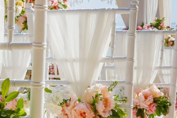 Weiße Hochzeitsstühle mit zarten Vorhängen und Blumenarrangements am Strand vor ruhigem Meer