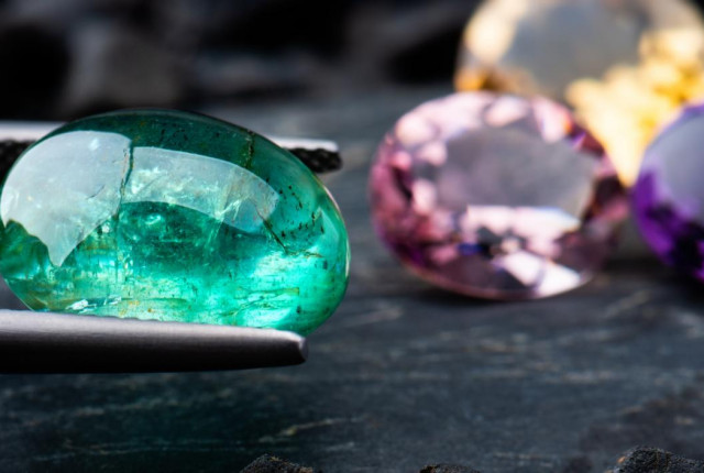 Oval emerald cabochon gemstone held in tweezers, with pink, yellow and purple birthstones behind