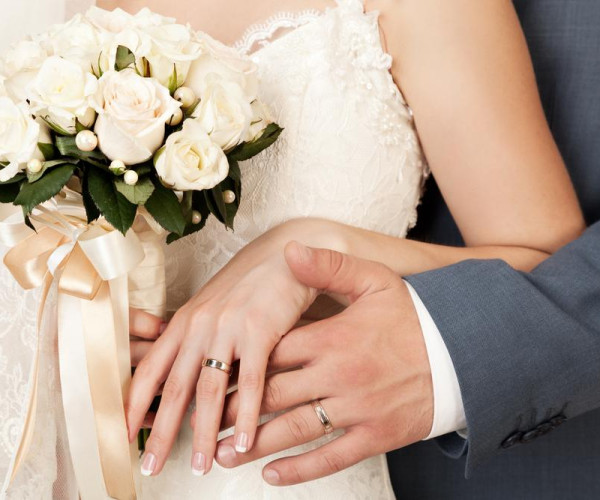 Bride and groom showing matching gold wedding bands, with the bride holding a white rose bouquet.