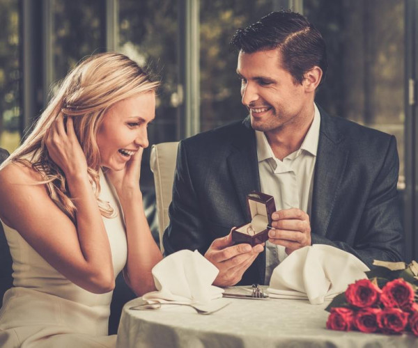 Man presenting a diamond engagement ring in a velvet box to a smiling woman at a romantic dinner table