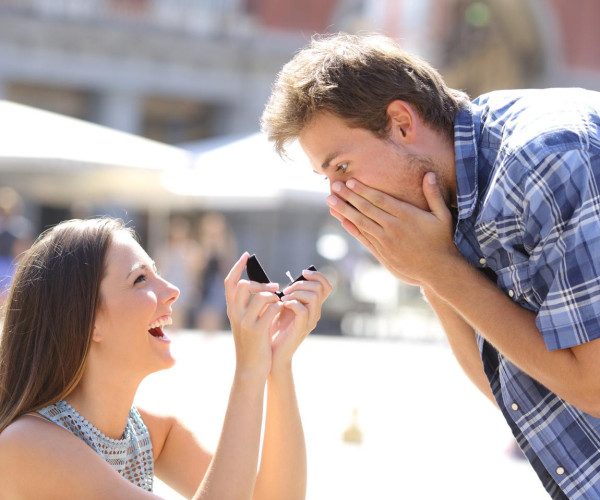 Woman proposing with a diamond ring in a black velvet box to a surprised man outdoors