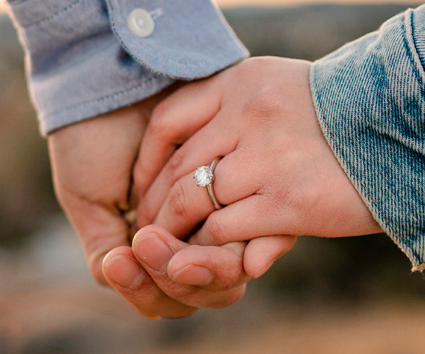 Hands holding in a romantic close-up, with a white gold solitaire diamond ring on the woman’s finger.