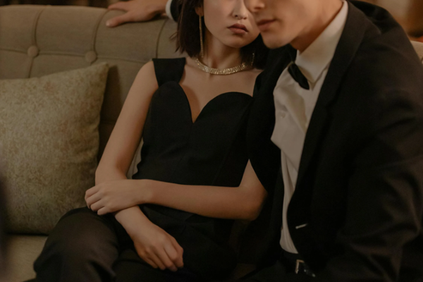A young couple in formal adorned in a chic black dress and dazzling diamond necklace and tailored black suit, gracefully positioned on a couch, exuding sophistication and a quiet sense of reflection.