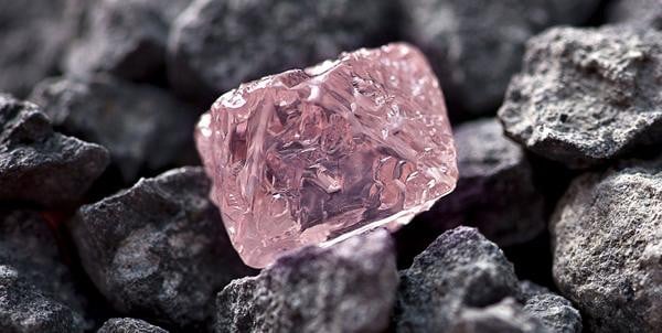 Rough pink diamond crystal in a close-up on dark rocks, showing natural faceted texture and translucent blush color