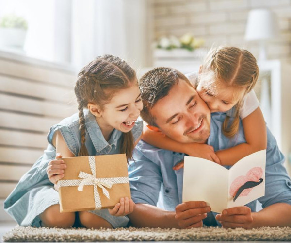 Padre con dos hijas leyendo una tarjeta y sosteniendo un regalo envuelto, escena familiar para el Día del Padre