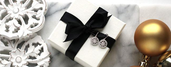 Un dessus de table en marbre avec des ornements de rosace blanche, une boîte blanche avec un cadeau de Noël, enveloppé d'un nœud noir avec deux boucles d'oreilles en diamant et des boules de Noël en or.