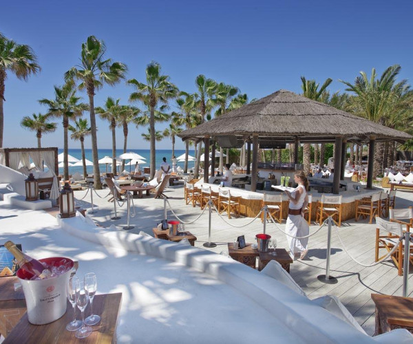 Lounge de plage tropicale à Saint-Tropez avec palmiers, bar en toit de chaume et tables blanches.