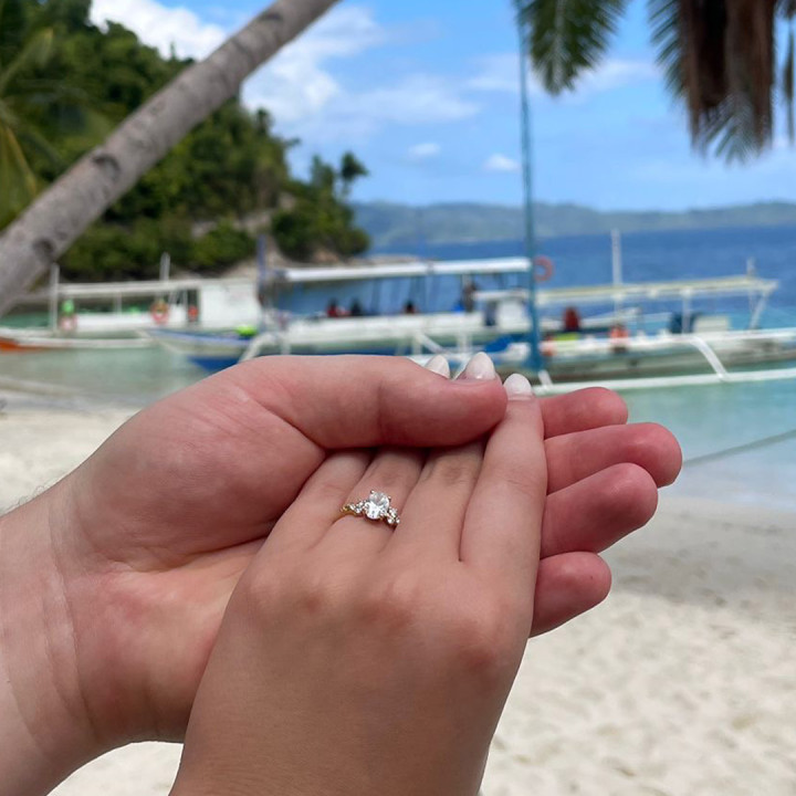海灘背景中，一對手輕握著手，女性手指戴著黃金鑲嵌的單顆圓形鑽石戒指，兩側點綴小鑽，優雅求婚戒設計