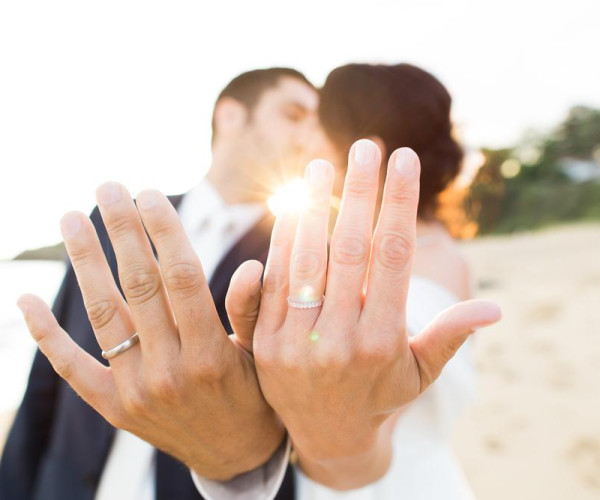 夕日のビーチで婚約・結婚指輪をつけた男女が左手を重ね、男性はシンプルなシルバーリング、女性はダイヤ付きの細身リングを見せている