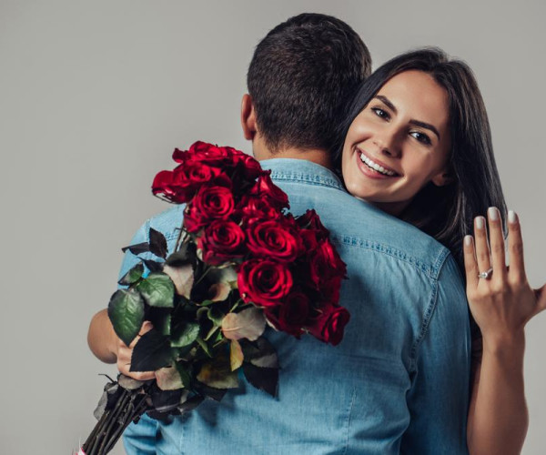 赤いバラの花束を持つ男性に抱きつき、婚約指輪を見せる笑顔の女性のプロポーズ風イメージ