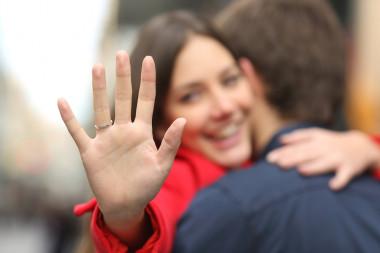 Verlobte Frau mit schmalem goldenen Ring ohne Stein an erhobener Hand, im Hintergrund Umarmung