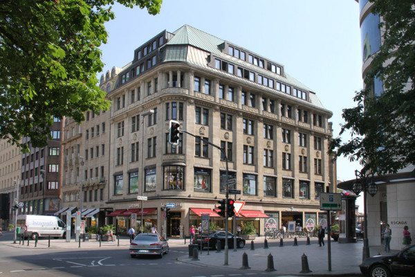 Historisches Sandsteingebäude an einer Straßenecke in Düsseldorf mit Ladenlokalen und grüner Kupferdachhaube.