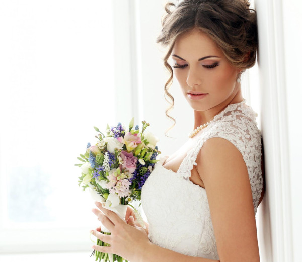 BAUNAT - Braut mit Blumenstrauß in weißem Hochzeitskleid lehnt an einer weißen Wand