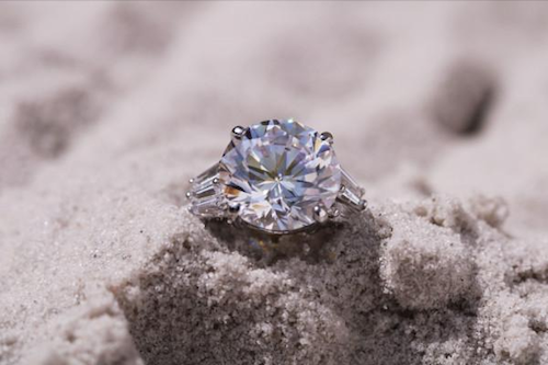 Gros plan d'une bague en diamant bleu clair avec une grosse gemme, enterrée dans du sable blanc.