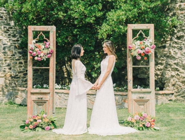 zwei Bräute lächeln bei einer gleichgeschlechtlichen Hochzeit im Vereinigten Königreich vor einem hölzernen Bogen, der von einer wunderschönen Außenkulisse umgeben ist