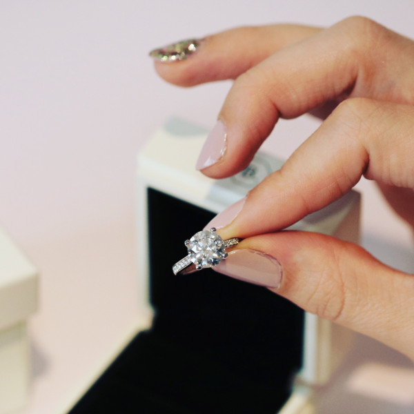  woman's hand holding a brilliant cut engagement ring in front of a white ring box