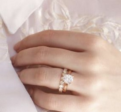 Een persoon die een witte geborduurde blouse draagt met zijn hand bij zijn kraag, met twee roodgouden ringen met een grote diamant en kleinere edelstenen.