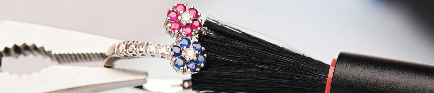 Gold ring met roze en blauwe edelstenen die klem zit tussen pincet en borstelharen Laat uw juwelen reinigen door een expert bij BAUNAT.