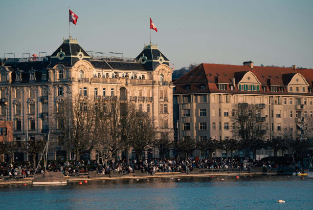 Panoramisch uitzicht op de Zürichse oever met historische gebouwen en Zwitserse vlaggen aan het meer.