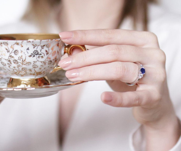 een vrouw met een kopje thee in de hand, die haar trilogie diamanten verlovingsring draagt met een saffier in het midden, geflankeerd door twee kleinere witte diamanten