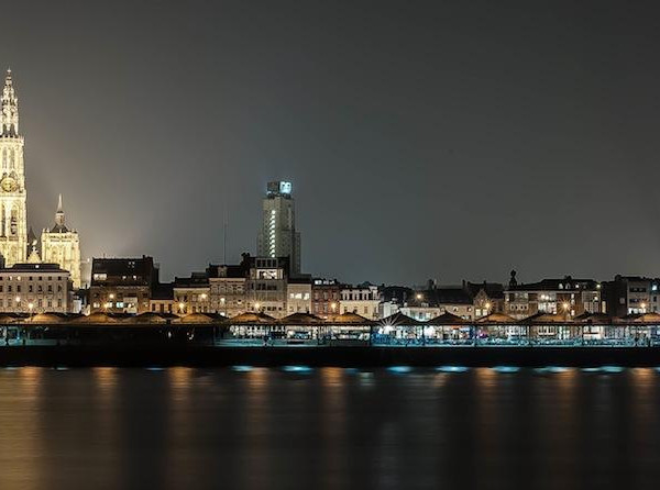 夜景城市天际线与河岸倒影，左侧高耸的灯光教堂塔楼、沿岸建筑和水面反光构成横向全景