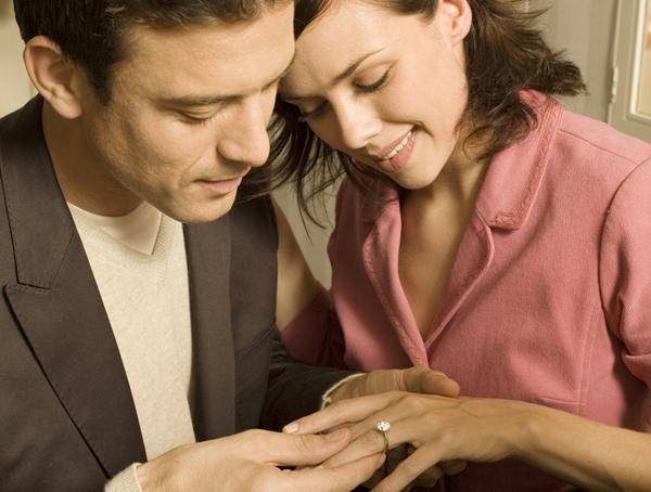 情侣近景展示白金单颗钻石订婚戒指，爪镶圆形主钻，佩戴在女性手指上，男士轻托其手，温馨浪漫场景
