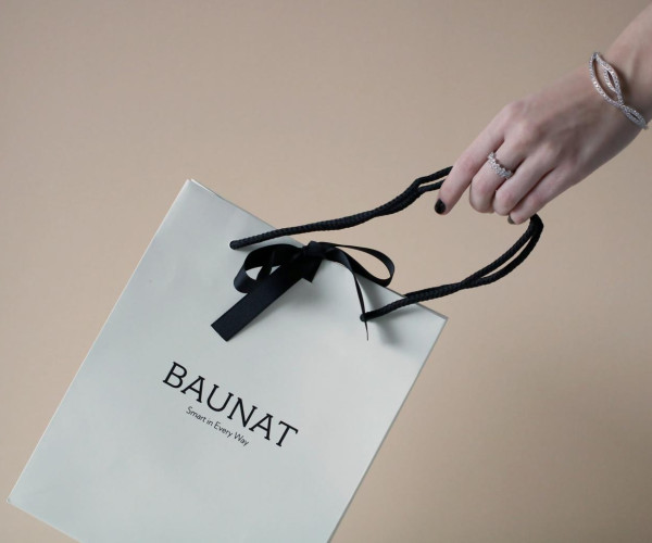 手持白色礼品袋，袋面印有 BAUNAT 标志，黑色丝带蝴蝶结与提绳，手腕和手指佩戴镶钻白金首饰