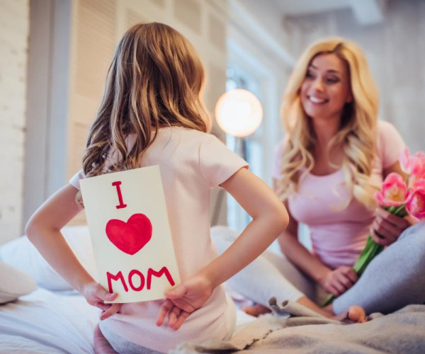 母亲节温馨场景：小女孩背后藏着写有“I love mom”的手工卡片，母亲手捧粉色花束坐在床上微笑相望